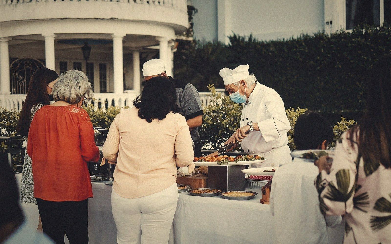 Chefs in masks serve guests at an outdoor buffet table beside a columned veranda